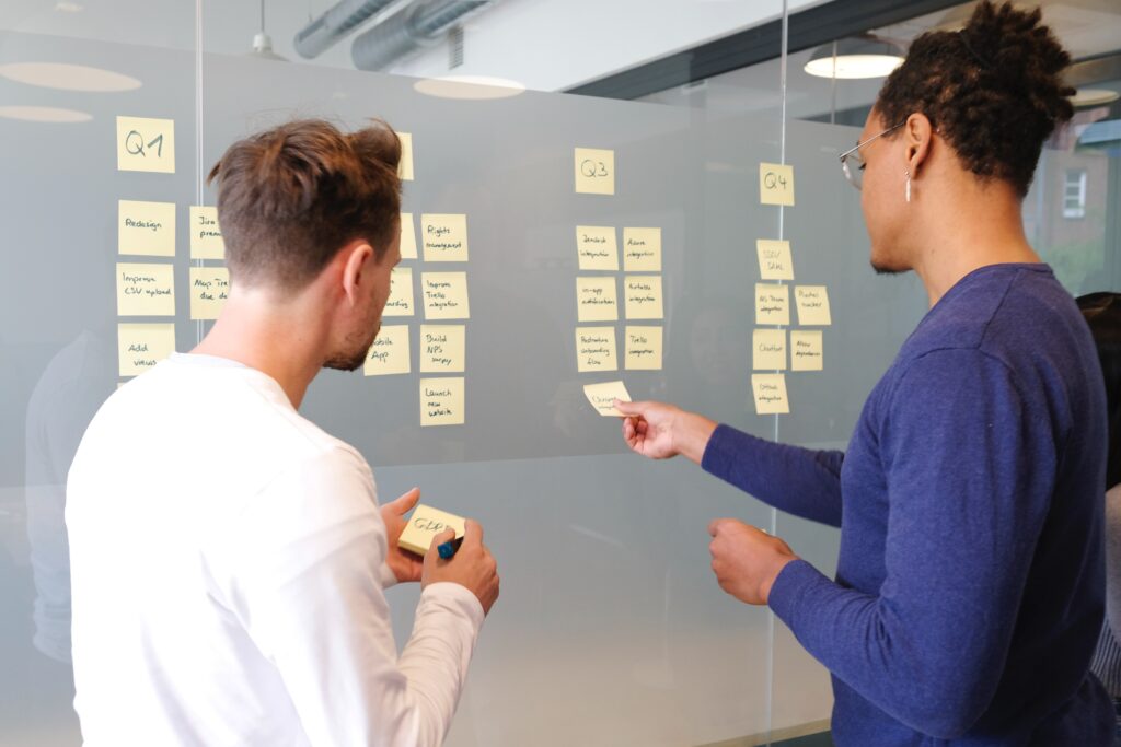 Two people stand before a glass wall, looking at several post-it notes displayed for review