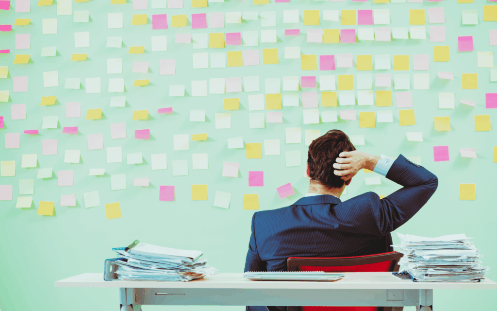 A man sits at a desk piled with papers. He is facing a wall of sticky notes while scratching the back of his head. 