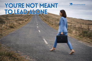 A professional woman in a powder blue business suit walks in profile along a quiet, empty road that stretches uphill into the distance. She carries a black briefcase in one hand and wears heels, moving with purpose under an overcast sky. The surrounding landscape is sparse and open, emphasizing the solitary journey ahead.