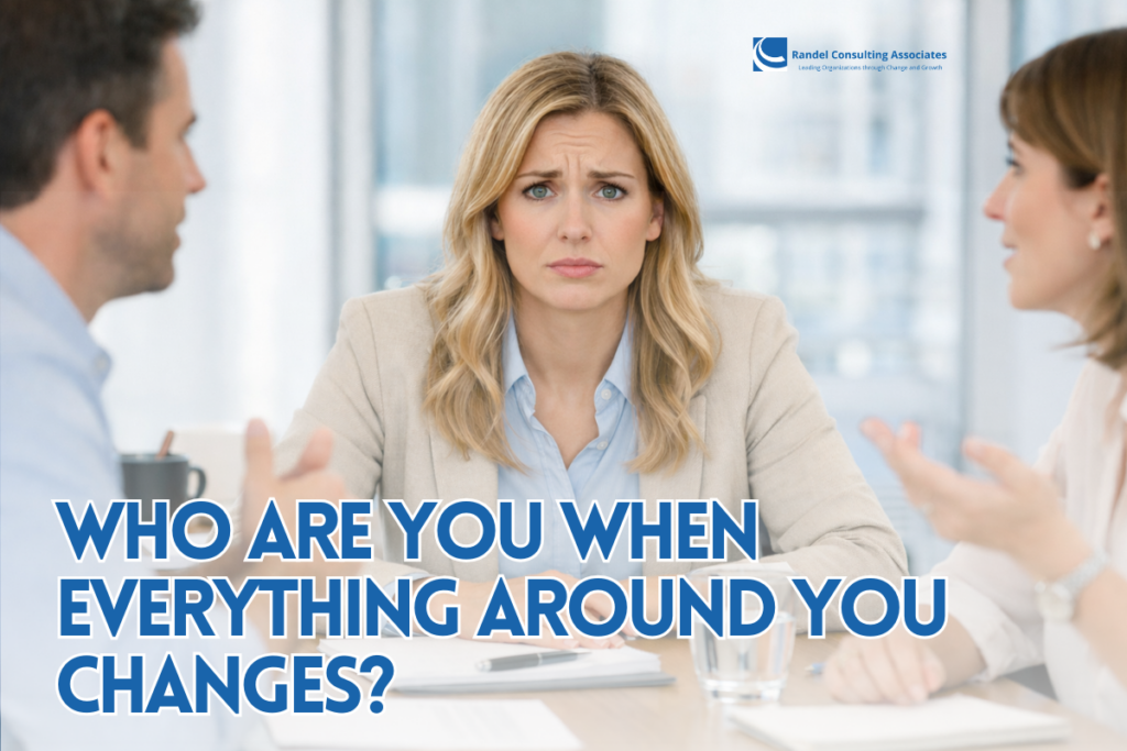 A professionally dressed woman sits at a boardroom table, facing forward with a concerned and slightly confused expression. She wears a light blazer over a blue shirt, with her hands resting on a notebook in front of her. A glass of water sits nearby. Two colleagues are partially visible on either side, gesturing as they speak, while the bright, modern office background is softly blurred, keeping the focus on her expression.