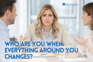 A professionally dressed woman sits at a boardroom table, facing forward with a concerned and slightly confused expression. She wears a light blazer over a blue shirt, with her hands resting on a notebook in front of her. A glass of water sits nearby. Two colleagues are partially visible on either side, gesturing as they speak, while the bright, modern office background is softly blurred, keeping the focus on her expression.