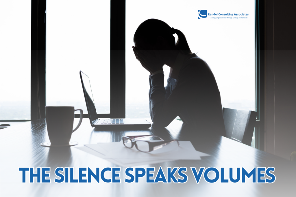 A silhouetted professional sits at a desk by a large window, head in hands, conveying stress or overwhelm. A laptop is open in front of them, with a coffee mug, glasses, and scattered papers on the table. The text “THE SILENCE SPEAKS VOLUMES” appears prominently at the bottom, with the Randel Consulting Associates logo in the top corner.