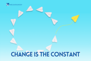 A series of white paper airplanes arranged in a circular pattern against a blue gradient background, symbolizing repetition and routine. One bright yellow paper airplane breaks away from the circle, flying upward along a dotted yellow path to represent change, growth, and transformation. The image includes the Randel Consulting Associates logo in the top left corner and the bold text “CHANGE IS THE CONSTANT” at the bottom.