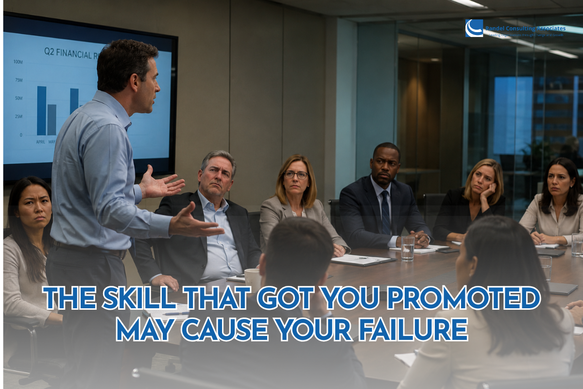 A business executive stands at the head of a conference room, presenting to a group of colleagues seated around a large meeting table. A financial report with bar charts is displayed on a screen behind him as the attendees watch with serious, attentive expressions. The image features the headline, "The Skill That Got You Promoted May Cause Your Failure," with the Randel Consulting Associates logo in the top-right corner, emphasizing the transition from individual contributor to effective leadership.